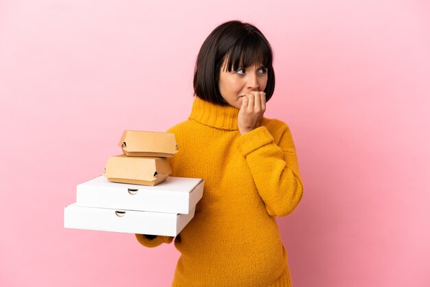 Pregnant woman holding pizzas and burgers isolated on pink background having doubts