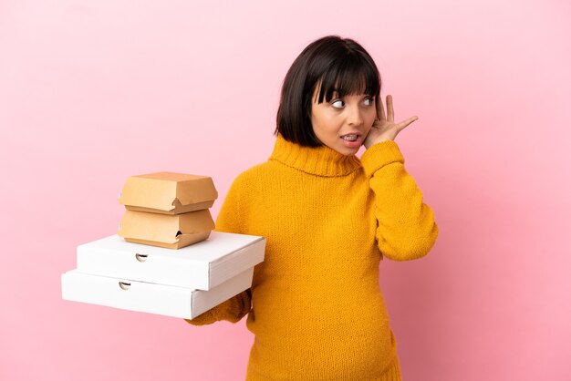 Pregnant woman holding pizzas and burgers isolated on pink background listening to something by putting hand on the ear