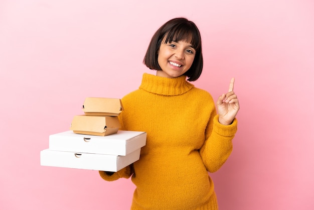 Pregnant woman holding pizzas and burgers isolated on pink background showing and lifting a finger in sign of the best