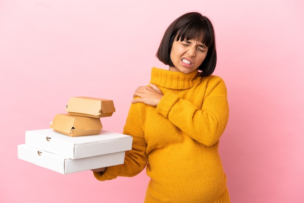 Pregnant woman holding pizzas and burgers isolated on pink background suffering from pain in shoulder for having made an effort