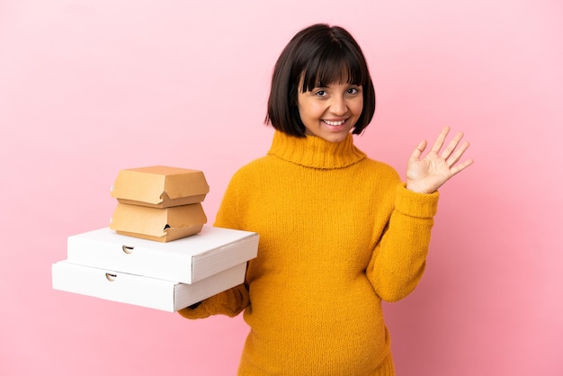 Foto donna incinta che tiene pizze e hamburger isolati su una superficie rosa che saluta con la mano con espressione felice