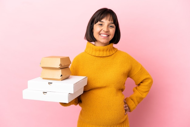 Pregnant woman holding pizzas and burgers isolated on pink wall posing with arms at hip and smiling