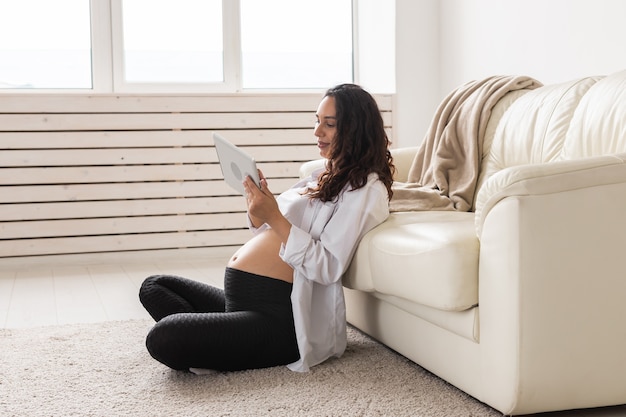 Pregnant woman holding tablet sitting on a carpet near a couch in the living room at home