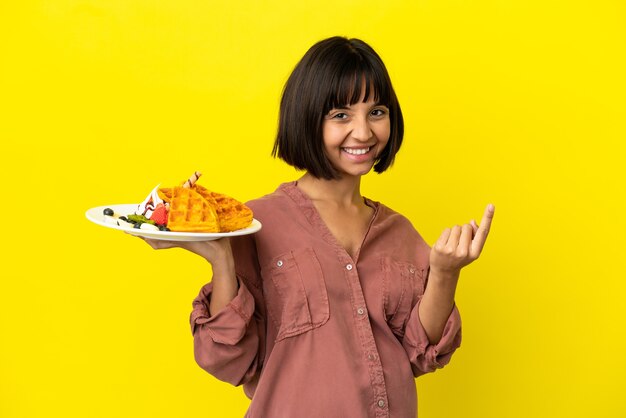 Pregnant woman holding waffles isolated on yellow background doing coming gesture