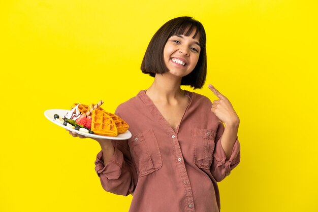 Foto donna incinta che tiene i waffle isolati su sfondo giallo che dà un gesto di pollice in alto