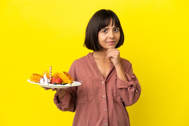 Pregnant woman holding waffles isolated on yellow background having doubts