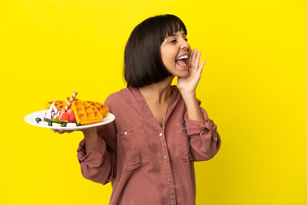 Pregnant woman holding waffles isolated on yellow background shouting with mouth wide open to the side