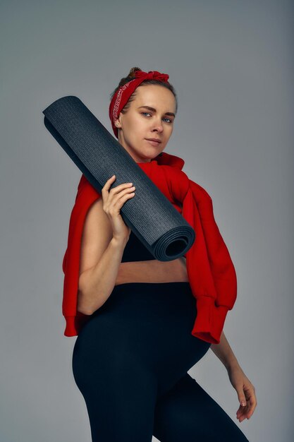 Pregnant woman holding yoga mat in hands going for sport while awaiting baby looking smiling at camera and touching her belly standing against gray wall