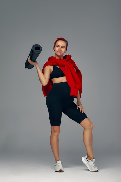 Pregnant woman holding yoga mat in hands going for sport while awaiting baby looking smiling at camera and touching her belly standing against gray wall