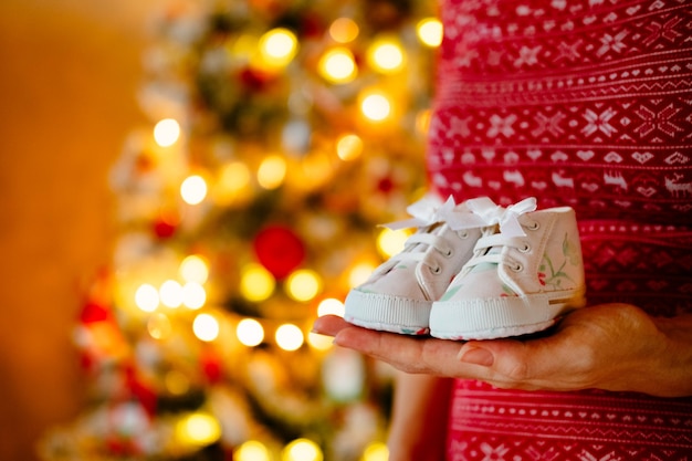 Pregnant woman holds cute little infants shoes in hands at christmas tree.