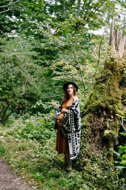 Pregnant woman hugs belly while standing near mossy tree near path in park