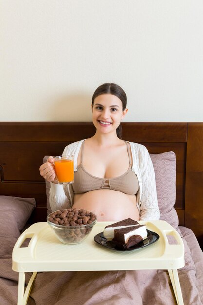 Pregnant woman is eating chocolate cereal balls piece of cake on breakfast tray and drinking juice