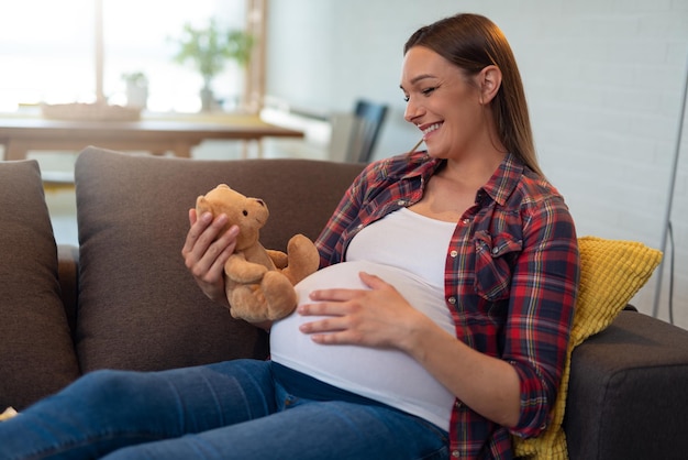 임신한 여자는 테디베어를 손에 들고 등을 대고 침대에 앉아 있습니다.