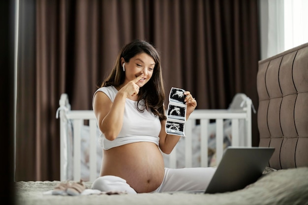 妊娠中の女性がベッドに座って、ラップトップで赤ちゃんの超音波写真を見せている