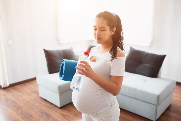 A pregnant woman is standing at home, she holds a yoga mat in her hands and drinks water after a workout. Close-up