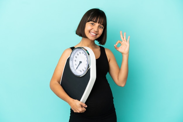 Pregnant woman over isolated background holding a weighing machine and doing OK sign