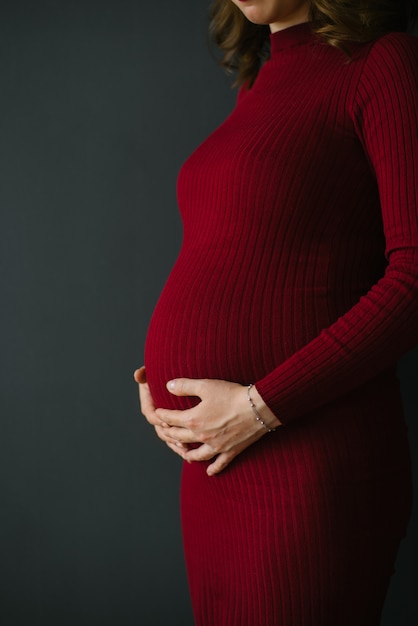 니트 부르고뉴 드레스에 임신 한 여자는 그녀의 뱃속에 그녀의 손을 보유하고있다. 아름답고 행복한 임신. 태어나지 않은 아이를 기다리는 중