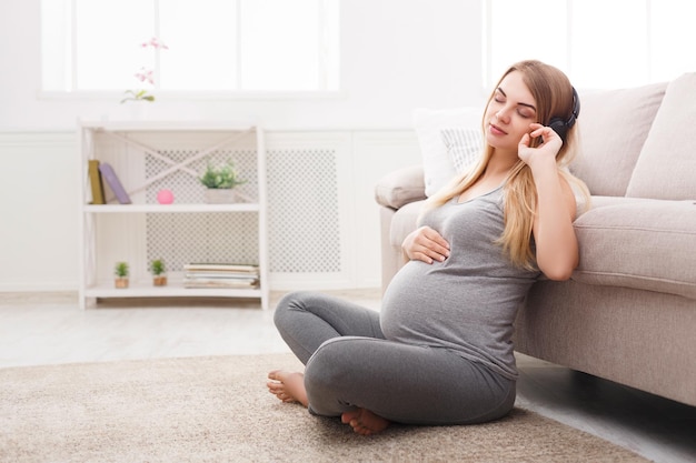 Pregnant woman listening to music in headphones. Young expectant blonde enjoying classical melody, copy space. Relax, leisure, pregnancy concept