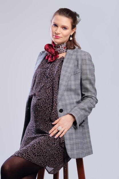 Pregnant woman in a loose dress and jacket sitting on chair on grey studio background