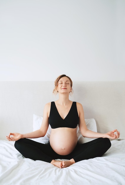 Pregnant woman in lotus pose doing meditation or breathing exercises for healthy pregnancy and preparing body for childbirth Young expectant mother practicing yoga at home