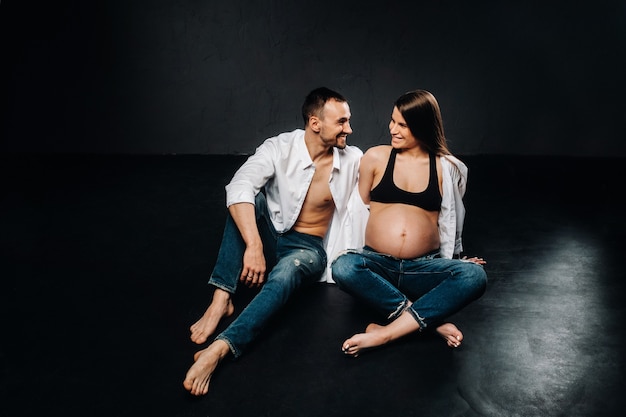 A pregnant woman and a man in a white shirt and jeans in a studio on a black background.