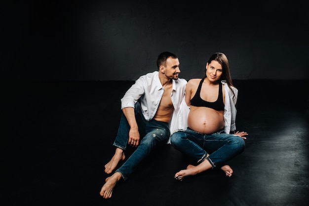 A pregnant woman and a man in a white shirt and jeans in a studio on a black background.