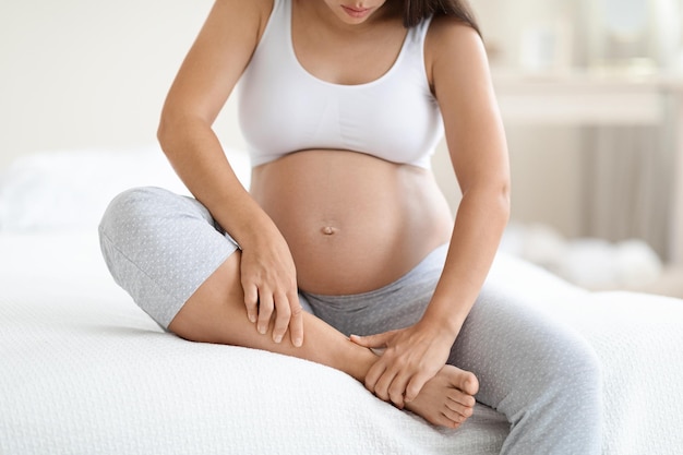 Pregnant woman massaging her swollen foot sitting on bed