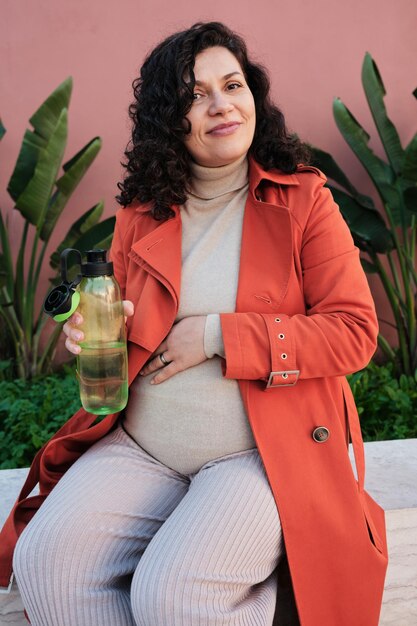 Photo pregnant woman in an orange coat on the street on a bench smiling drinking water