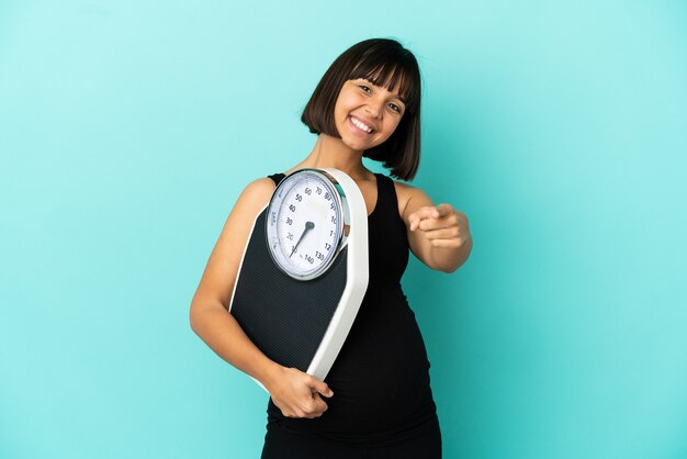 Беременная женщина на изолированном фоне держит весы и указывает вперед