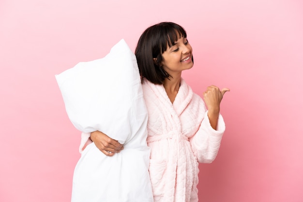 Pregnant woman in pajamas isolated on pink wall pointing to the side to present a product