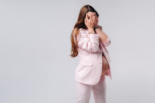 A pregnant woman in a pink suit close-up on a gray background covers her face with her hands