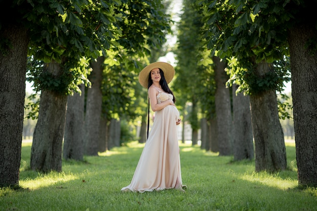Беременная женщина позирует в бежевом платье на фоне зеленых деревьев