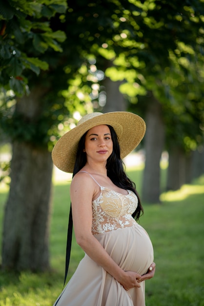 Pregnant woman posing in a beige dress on green trees.