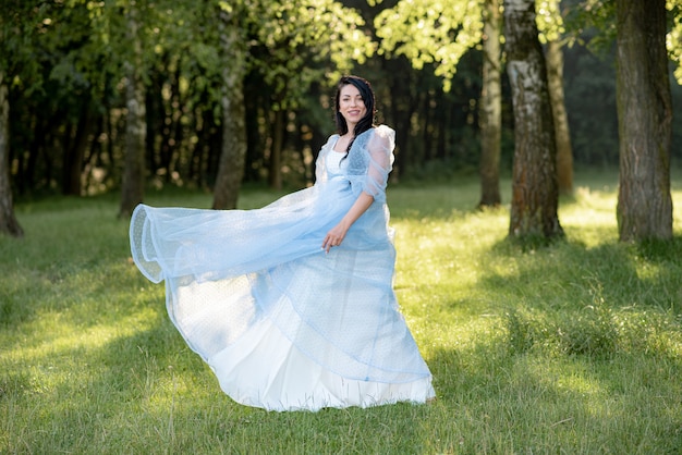 妊娠中の女性が緑の木々の背景に青いドレスでポーズします。