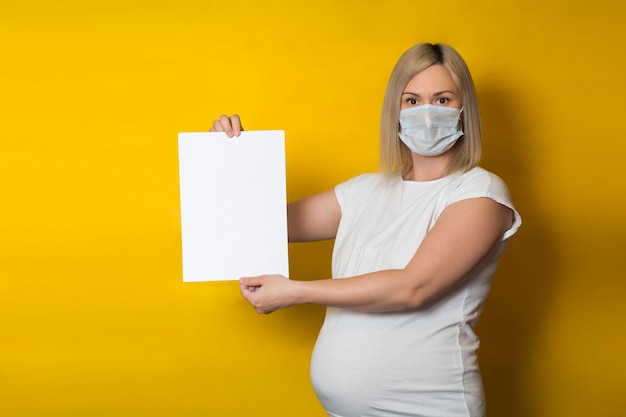 Photo a pregnant woman in a protective mask holds a blank vertical sheet of paper on a yellow wall