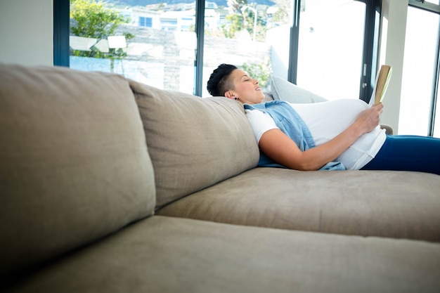 Pregnant woman reading a book while lying on sofa in living room