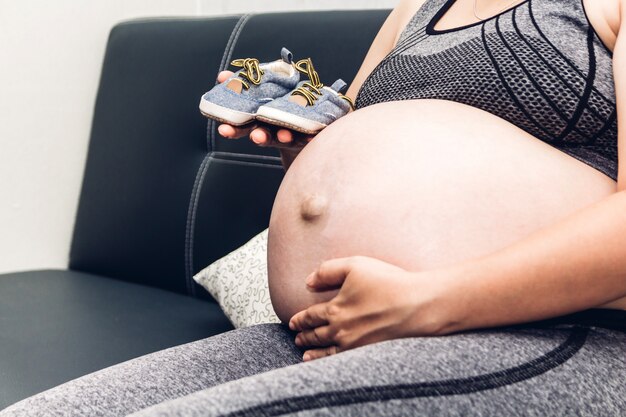 Pregnant woman relaxing and holding baby shoes on her belly for unborn baby on couch