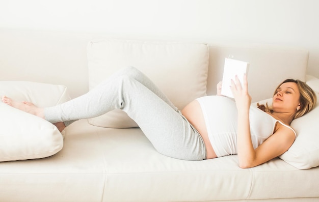 Pregnant woman resting at home and reading book on sofa