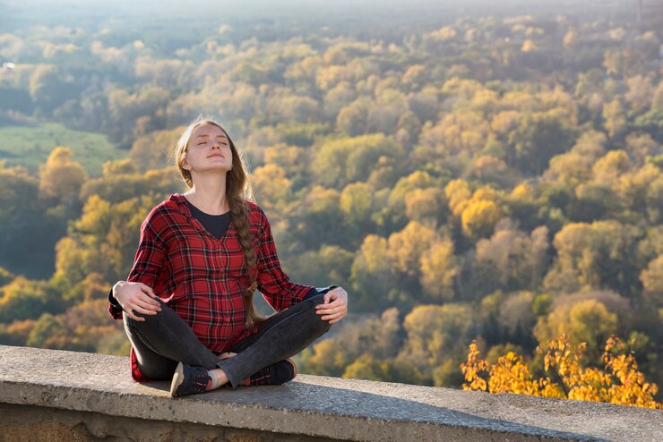 Pregnant woman sits on a hill with her eyes closed. 