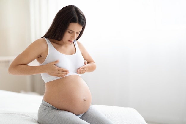 Pregnant woman sitting on bed having painful feelings in breast