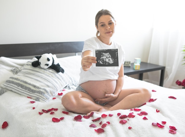 Pregnant woman sitting on a bed holding an Xray image of her babyThe concept of pregnancy motherhood and prenatal care Mom with a new life