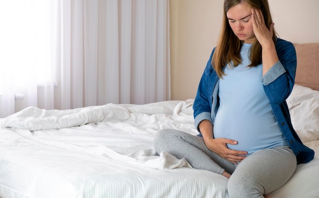 La donna incinta seduta a letto a casa sentendosi male toccare la pancia e la testa soffre di mal di testa o emicrania problemi sanitari e di gravidanza