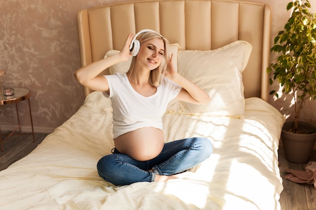 Pregnant woman sitting on bed at home and listening music in headphones