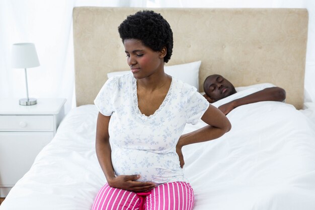 Pregnant woman sitting on bed while husband sleeping behind