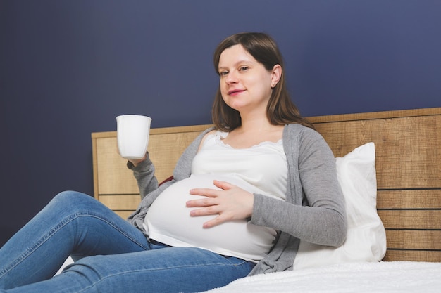 Pregnant woman sitting in bed with a cup of beverage with one hand on her belly