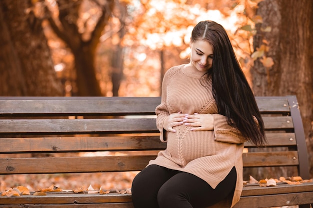 Pregnant woman sitting on the bench in autumn city park forest, holding stroking her round belly