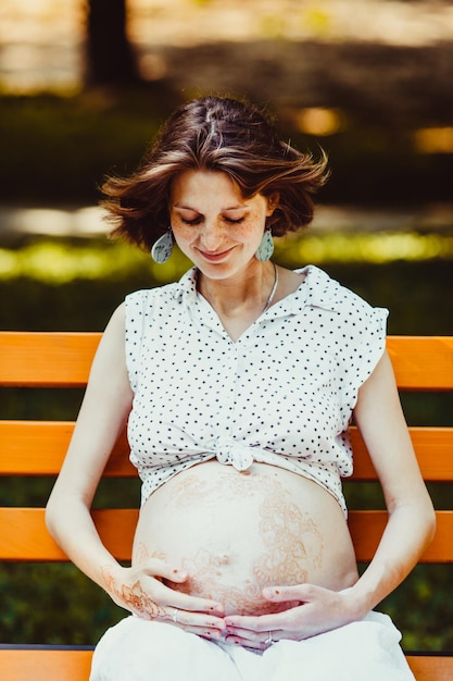 Pregnant Woman sitting on the bench in the park. Mehndi painting on the arm and belly