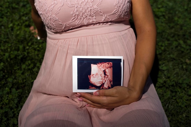 妊娠中の女性が彼女の子宮で赤ちゃんのイメージ 3 D 超音波を保持している芝生の上の公園に座っています。