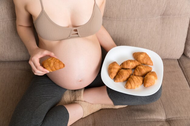 Pregnant woman sitting on the sofa eating croissants