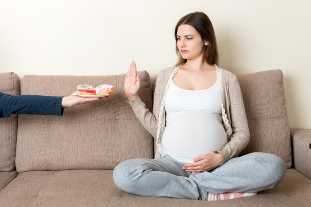 Pregnant woman sitting on the sofa rejects to eat junk food such as donuts and makes no gesture Healthy diet for future mother concept